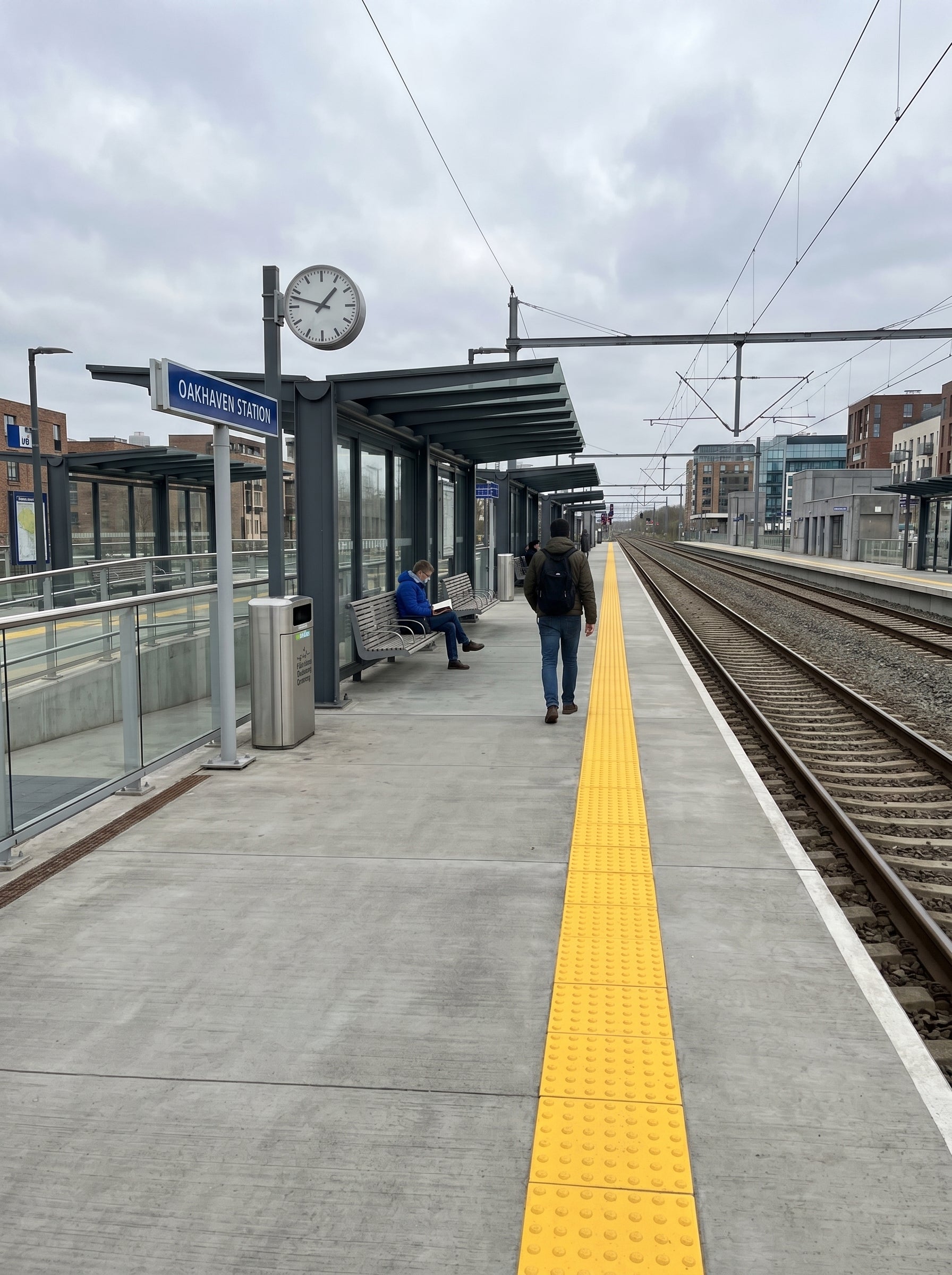 Alpha Vision - Train platform with a clock, benches, and a person walking on a cloudy day.